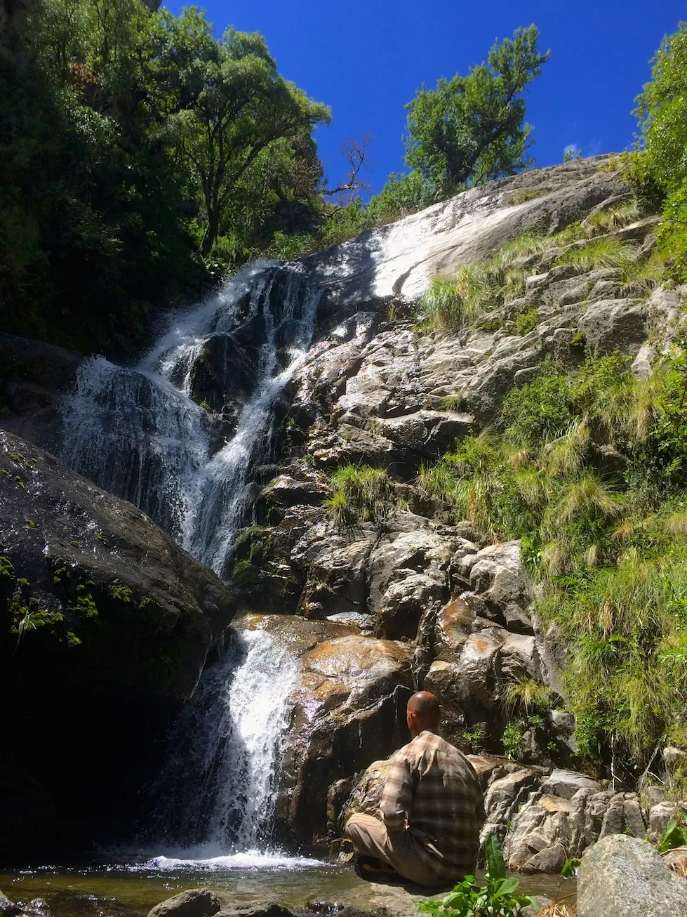 Una cascada alta cayendo por la pared de roca, con una persona sentada al pie.