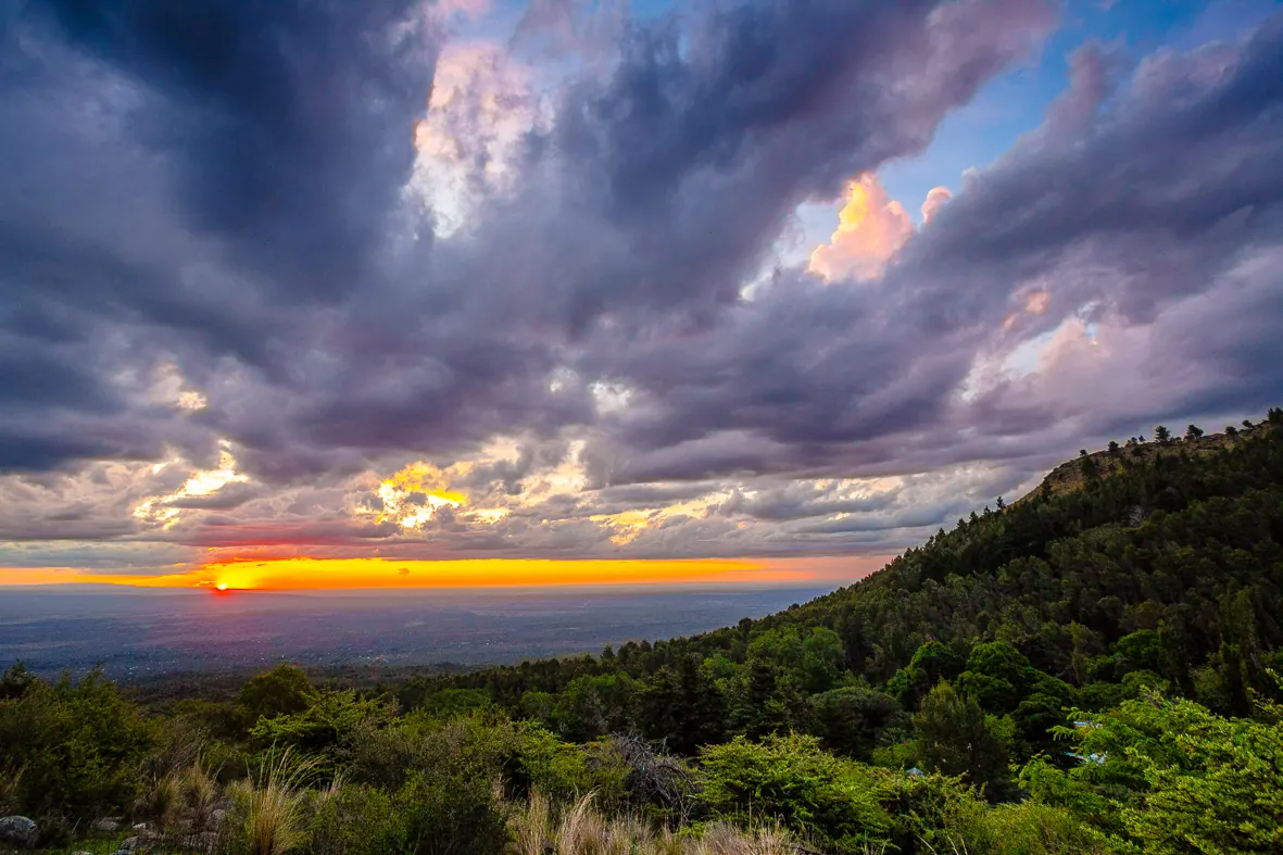 Atardecer desde el filo, con nubes cargadas y el sol bajo sobre el llano.