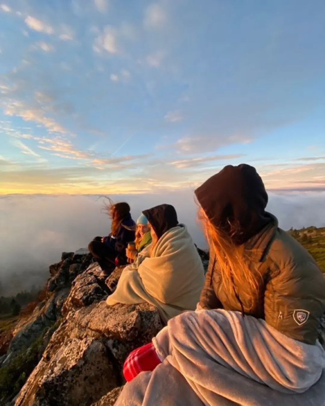 Gente envuelta en mantas mirando el amanecer por encima del mar de nubes.