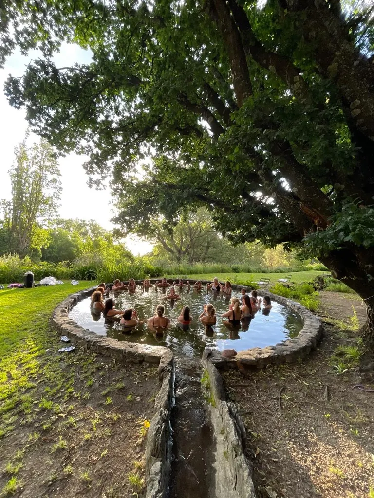 El estanque redondo de piedra bajo un árbol grande, con gente adentro del agua al atardecer.