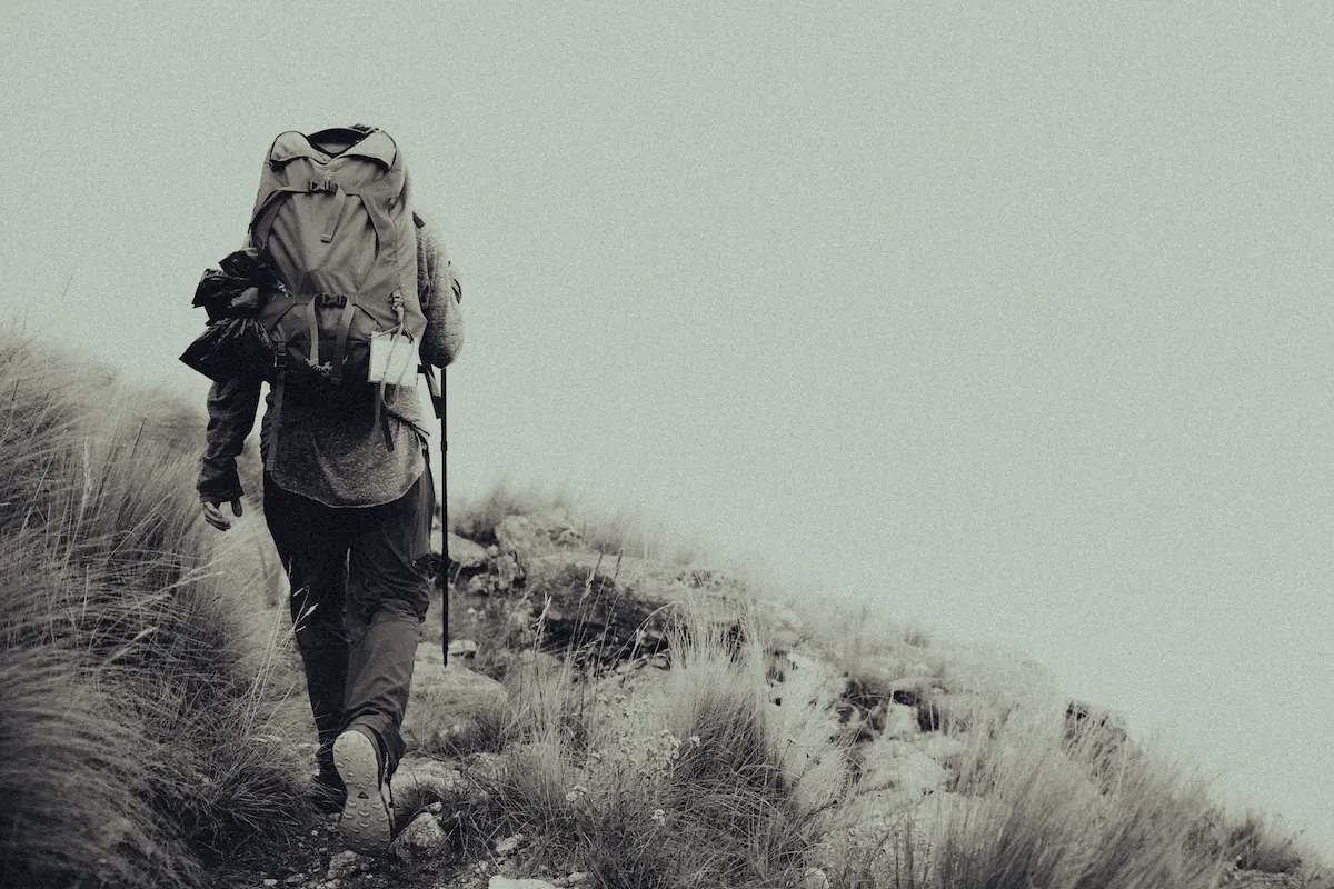 Un caminante con mochila grande subiendo por el pastizal, entre la niebla.