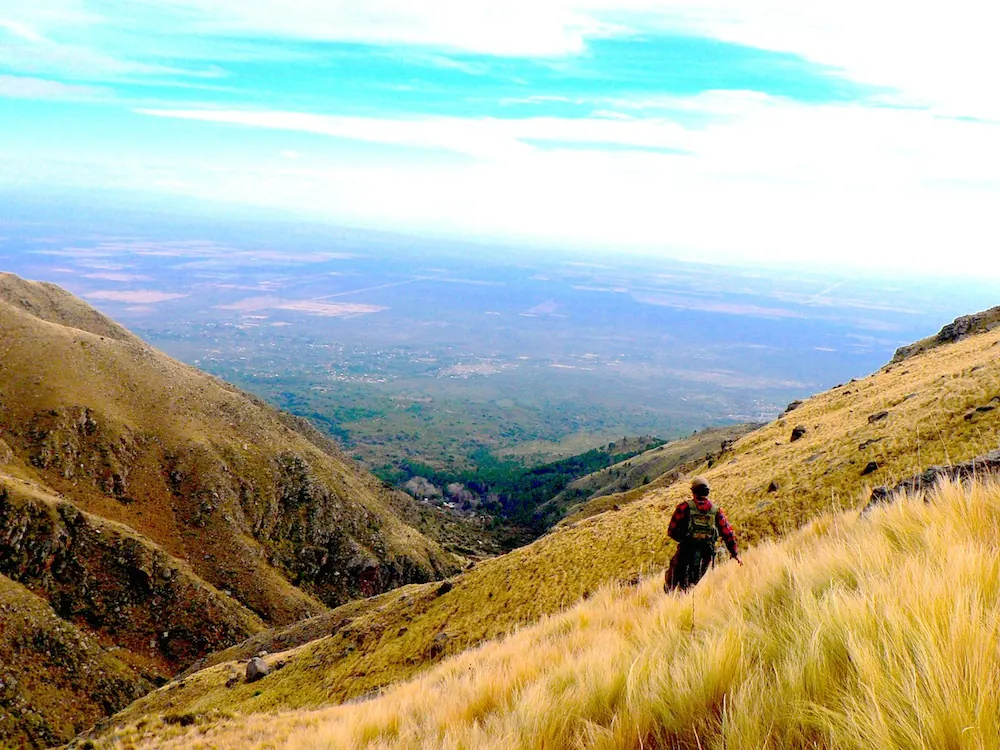 Una persona bajando por el sendero entre pastizal de altura, con el valle abierto al fondo.
