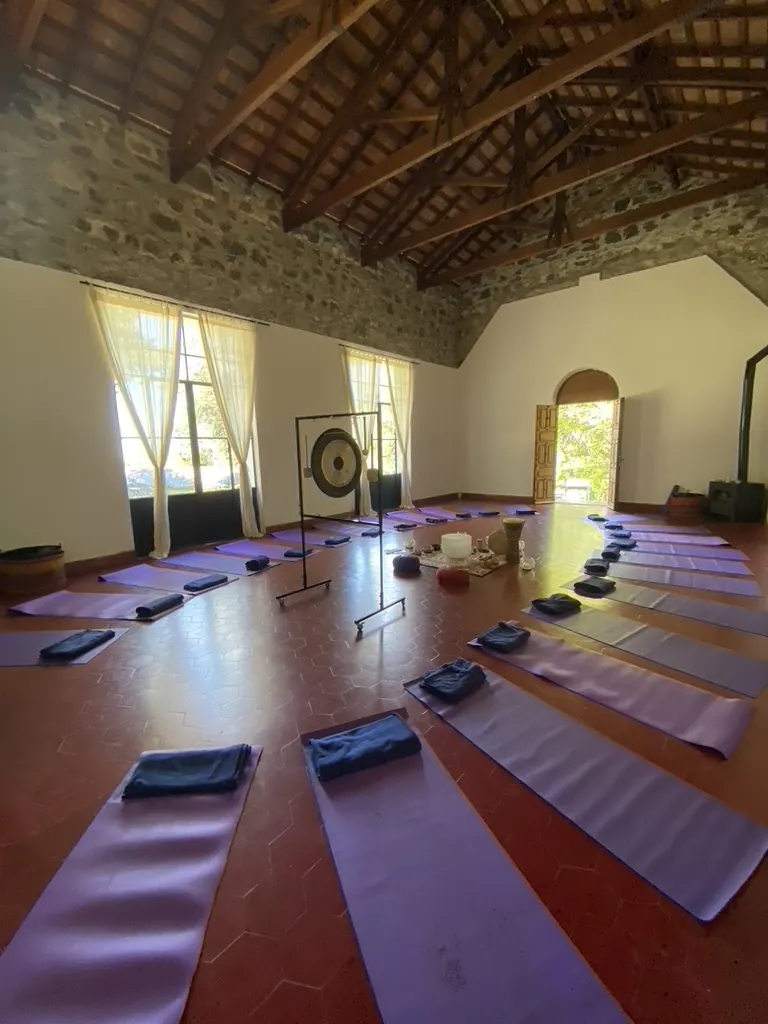 Un salón con techo de vigas y piso rojo, con colchonetas y un gong.