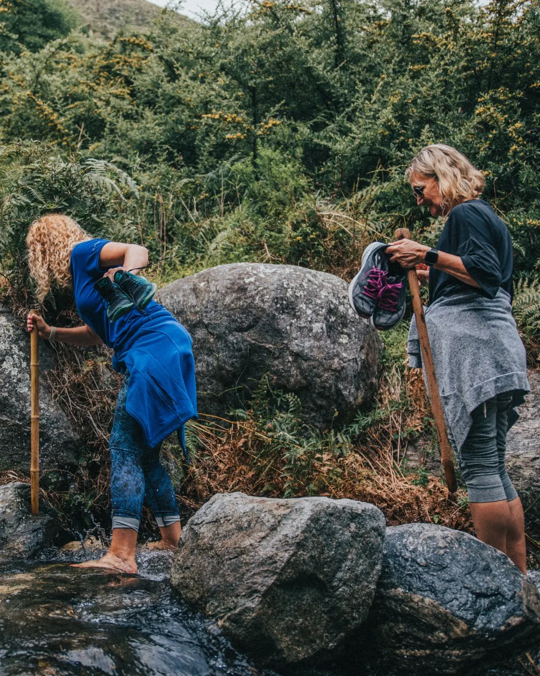 Dos personas cruzando el arroyo descalzas, con bastones.