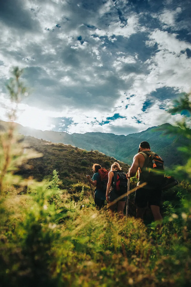 Grupo subiendo por el pastizal con mochilas, bajo un cielo cargado.
