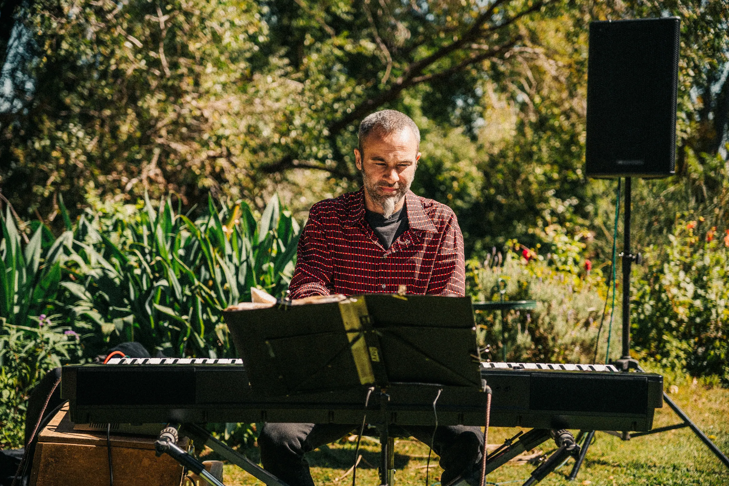 Un músico tocando el teclado en el jardín, entre las plantas, con un parlante al costado.