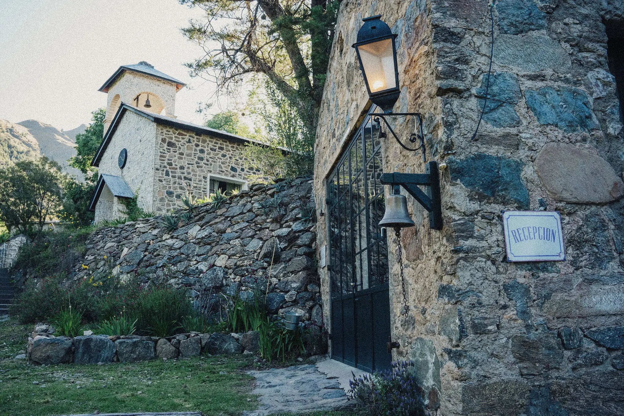 La campana de bronce en la pared de piedra, con el oratorio detras.