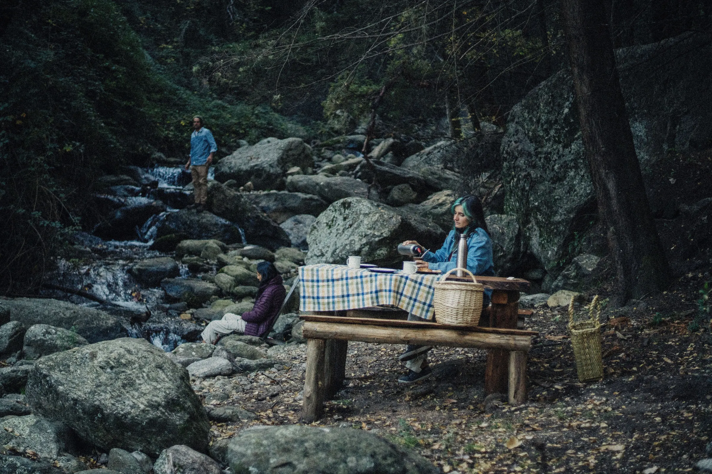 Una mesa de madera junto al arroyo, con mantel y canasto, y gente sentada alrededor entre las rocas.