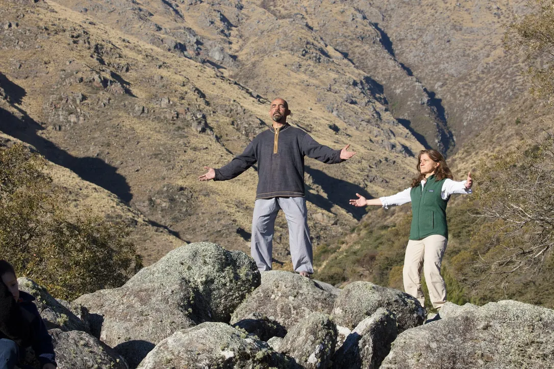 Un hombre y una mujer de pie sobre las rocas con los brazos abiertos y los ojos cerrados, frente a la ladera dorada de la sierra.