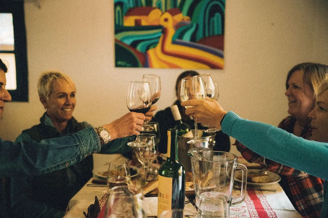 Cuatro personas chocando las copas de vino sobre la mesa puesta, riéndose, con un cuadro de colores en la pared.