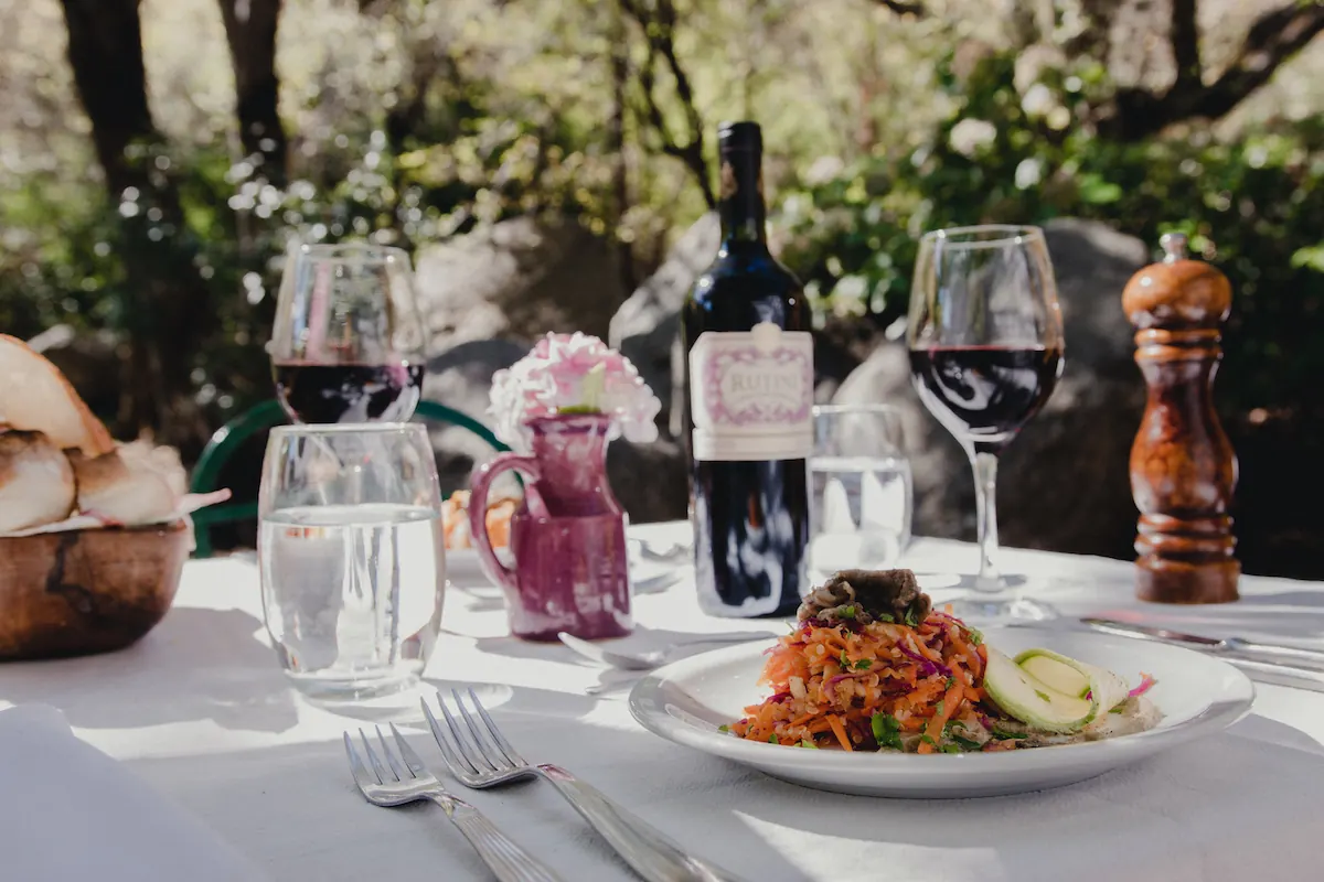Una mesa servida al aire libre con mantel blanco, vino y un plato de pastas.