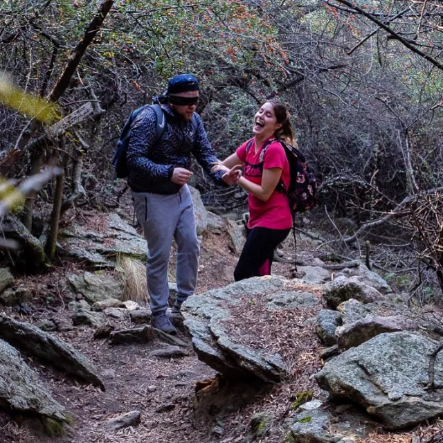 Una persona con los ojos vendados avanza por el sendero de piedra, guiada de la mano por otra que se ríe.