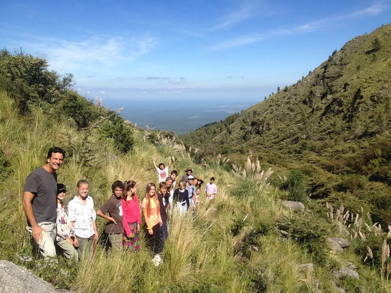 Un grupo de chicos y adolescentes con un guía, parados en un sendero entre el pastizal, con el llano de Traslasierra al fondo.