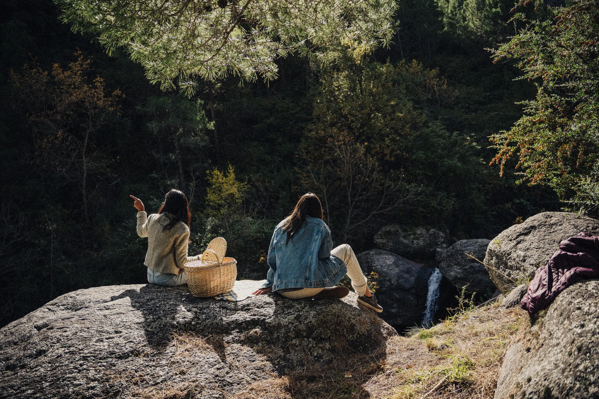 Dos personas sentadas sobre las rocas grandes del arroyo con un canasto de mimbre, una señalando hacia el bosque.