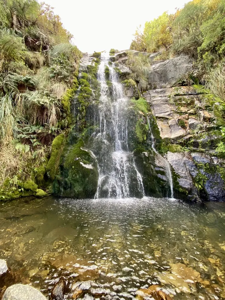 La cascada cayendo por la pared de roca cubierta de musgo, con el pozón de agua clara al pie.