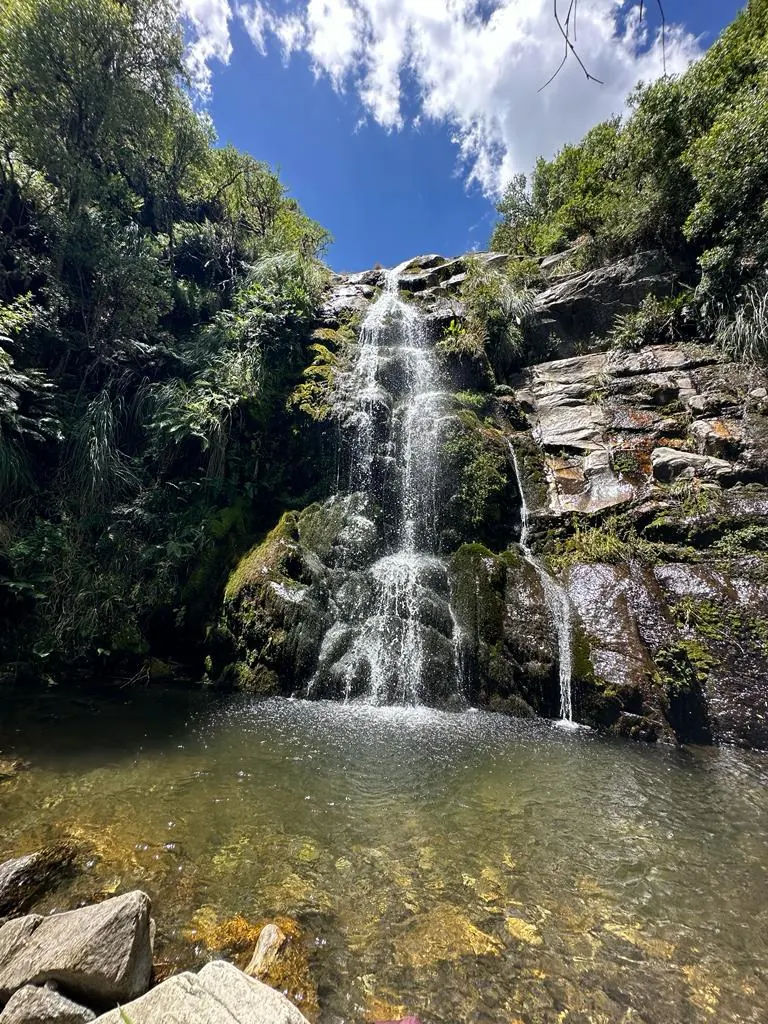 La cascada cayendo por la pared de roca cubierta de musgo hasta el pozón de agua clara, con el cielo azul arriba.