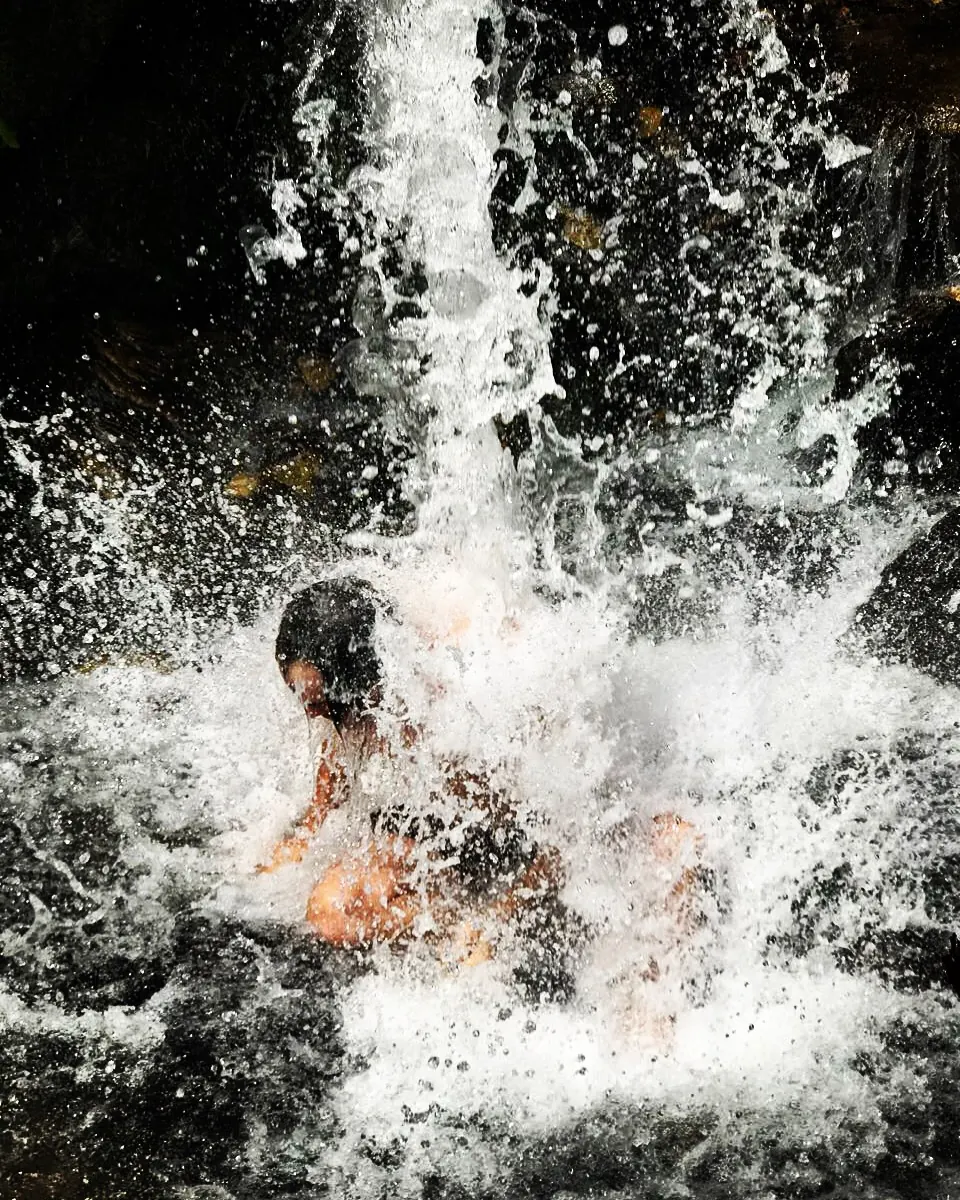 Un chico agachado bajo el chorro de la cascada, envuelto en el agua que salpica.