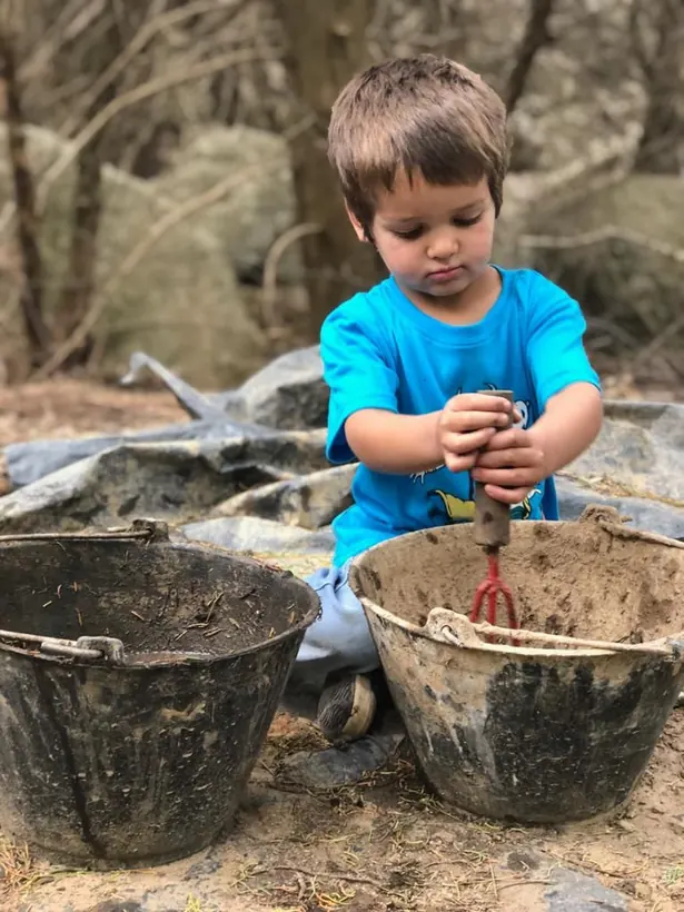 Un chico muy chico sentado en el suelo, llenando un balde de tierra con una pala de jardín.