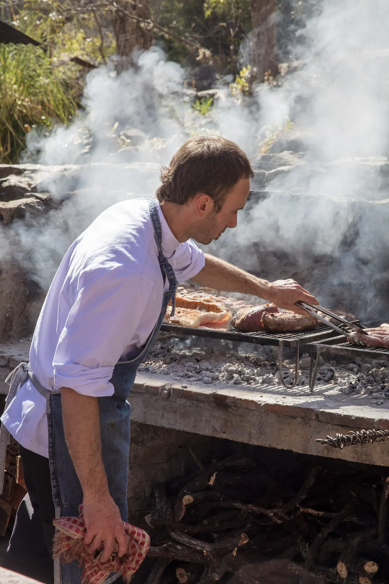 El cocinero de delantal dando vuelta la carne en la parrilla de piedra, entre el humo, con el monte detrás.