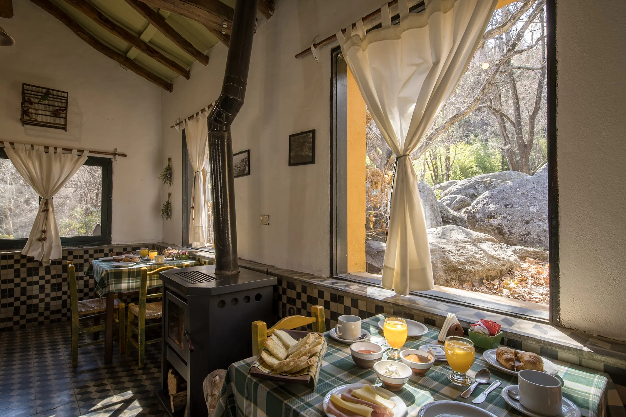 Un comedor chico con mesas puestas para el desayuno, una salamandra en el medio y ventanales con cortinas blancas que dan a las rocas y el bosque.