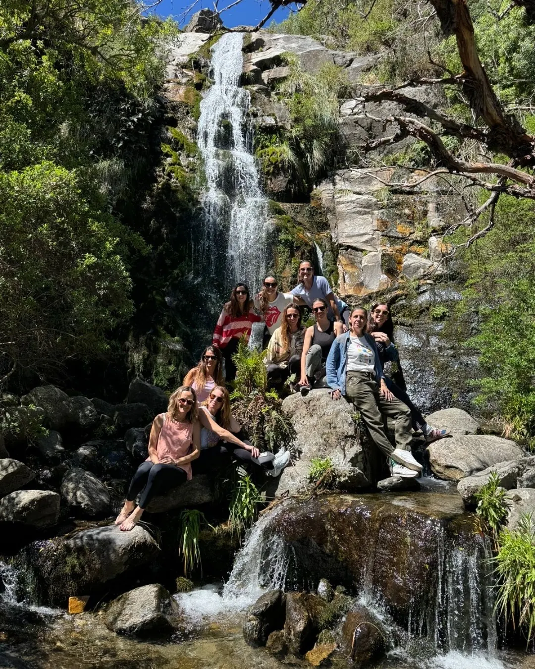 Un grupo sentado en las rocas al pie de la cascada, entre el agua y la vegetación.