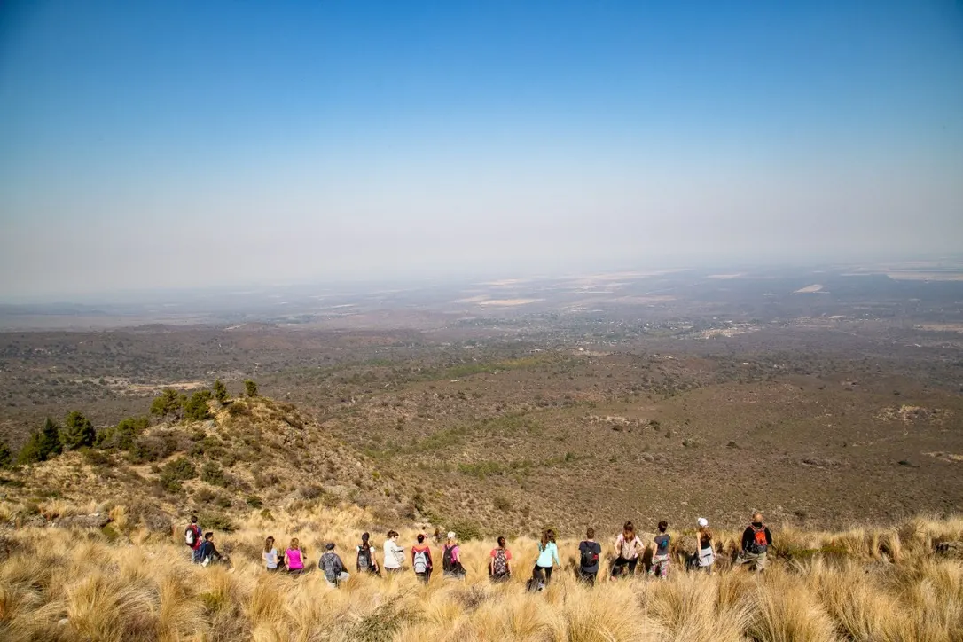 Una fila de caminantes de espaldas entre los pastizales del filo, mirando el llano de Traslasierra que se pierde en la bruma.