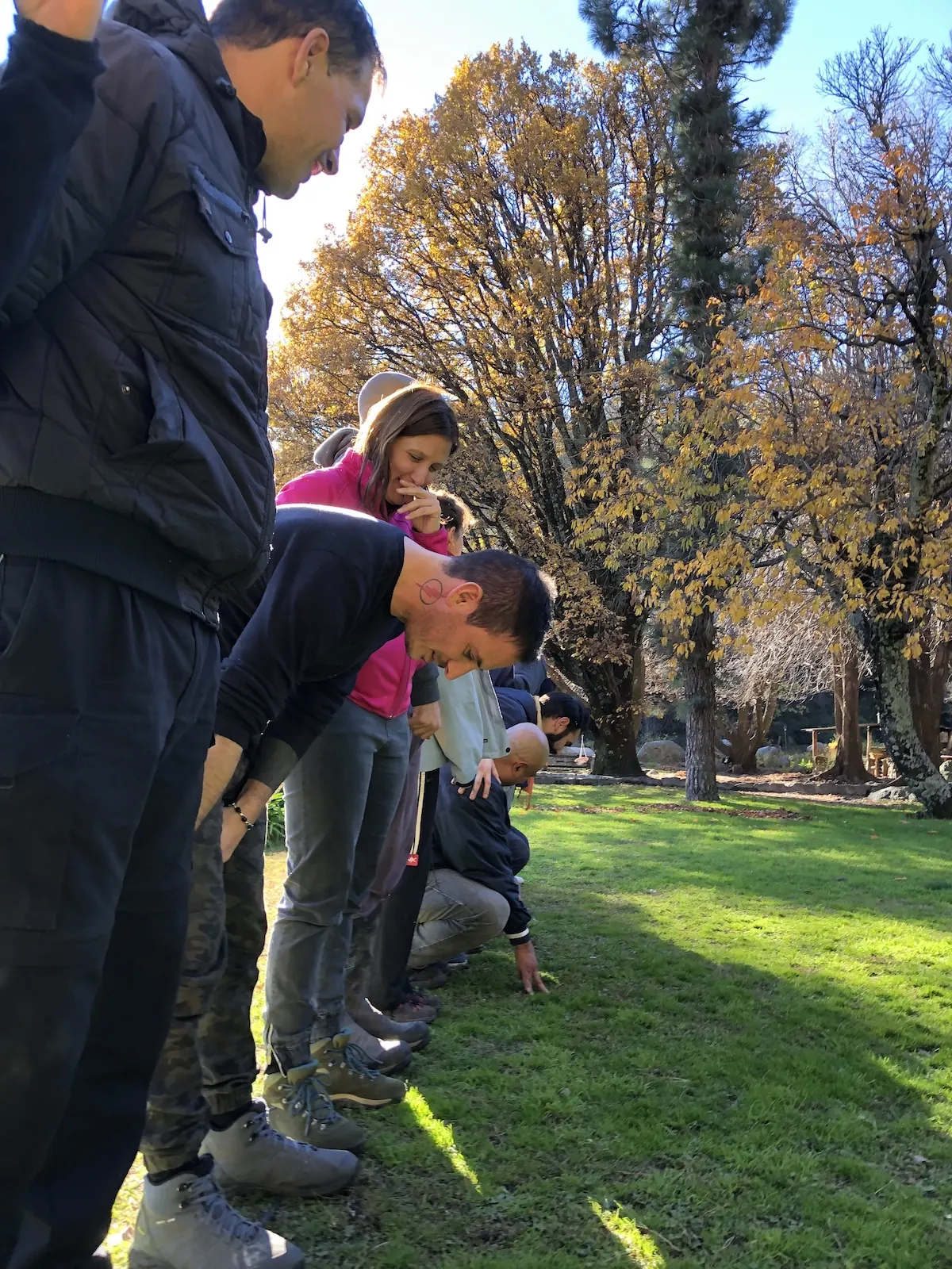 Una fila de personas paradas en el parque, en otoño, inclinadas mirando el pasto.