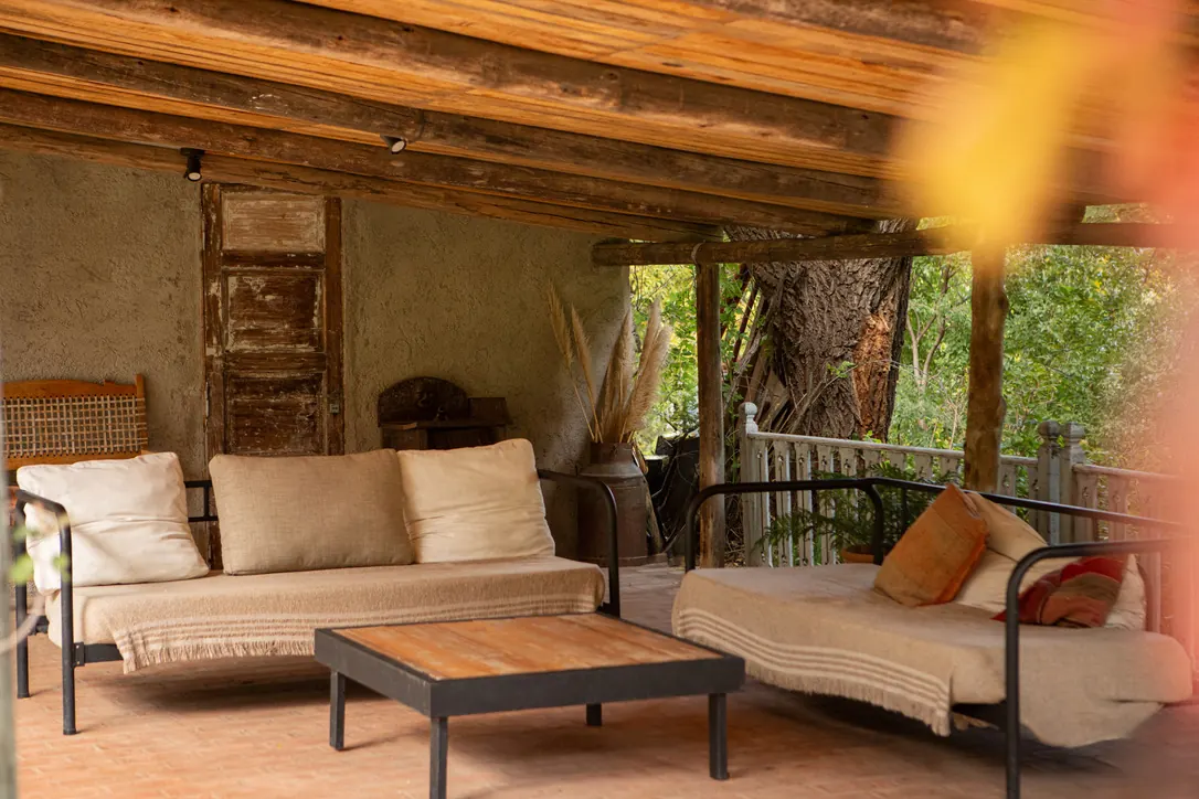 La galería techada con vigas de madera, dos sillones bajos con mantas claras y una mesa ratona, abierta al bosque por una baranda de troncos.