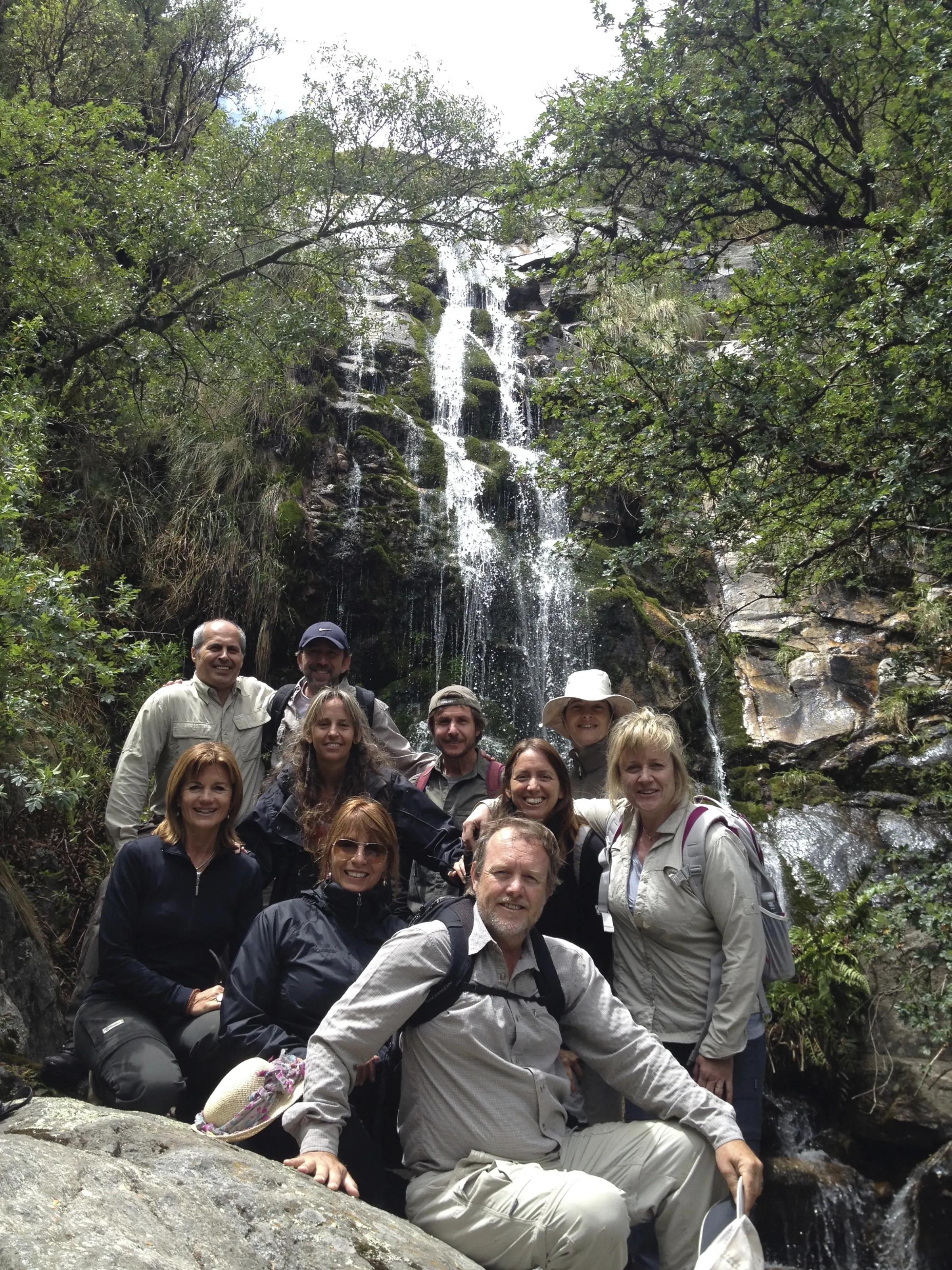Un grupo de diez personas con mochilas posando sobre las rocas al pie de la cascada, entre la vegetación.