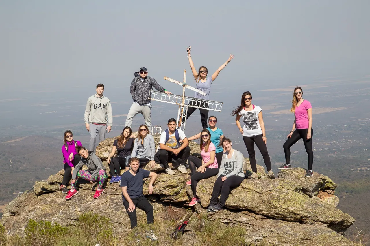 Un equipo de quince personas posando sobre las rocas de una cumbre, alrededor de una cruz de hierro, con el valle abajo.
