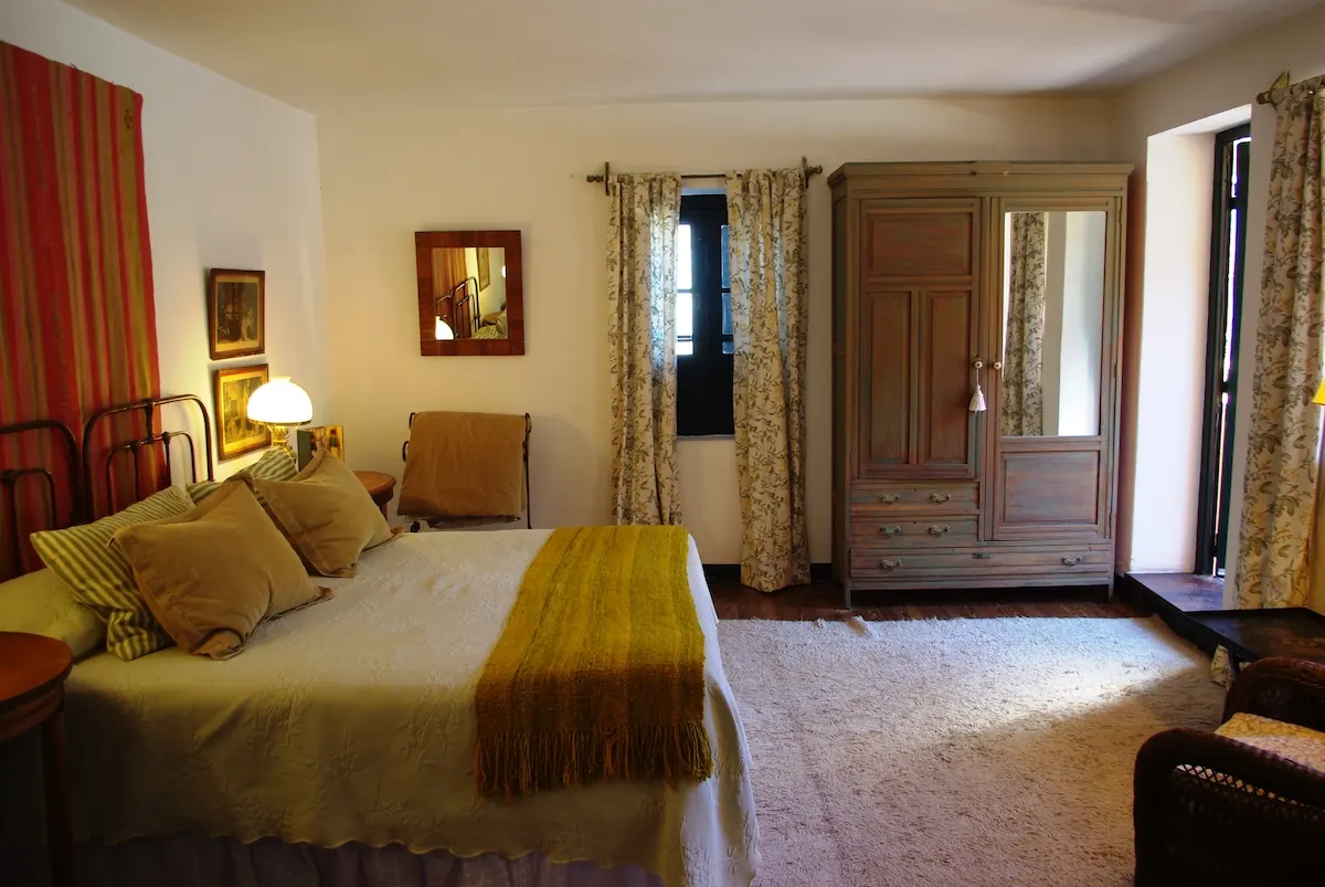 Habitación con cama matrimonial de acolchado dorado, ropero de madera con espejo y cortinas floreadas.