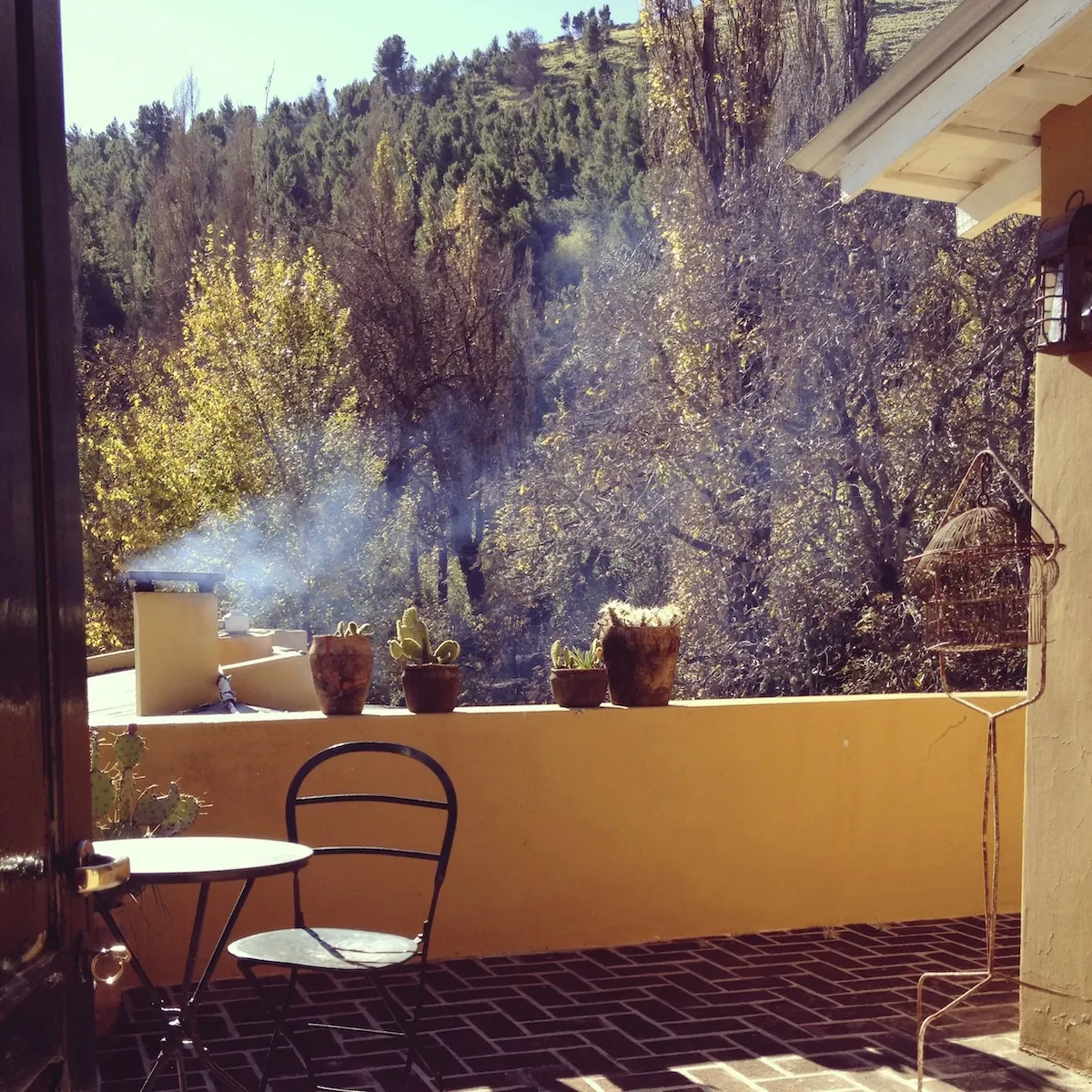 Terraza con macetas sobre el muro y una mesa con dos sillas, mirando a los árboles de la quebrada.