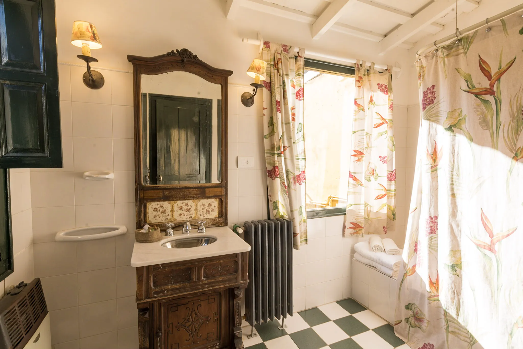 Baño de piso ajedrezado con mueble de madera antiguo, espejo y cortina floreada.