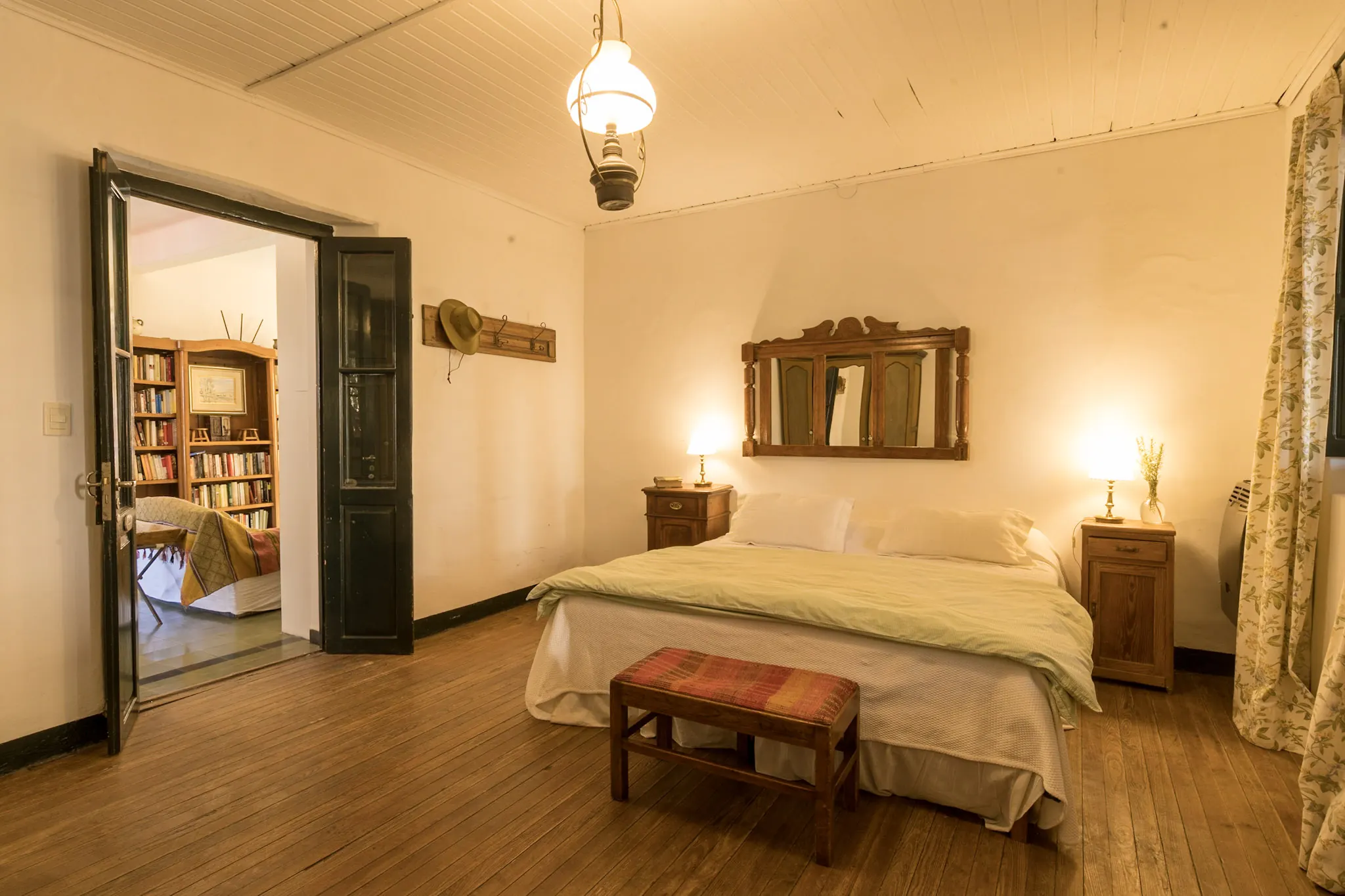 Habitación con cama matrimonial y una puerta doble abierta que deja ver la sala de lectura con biblioteca.