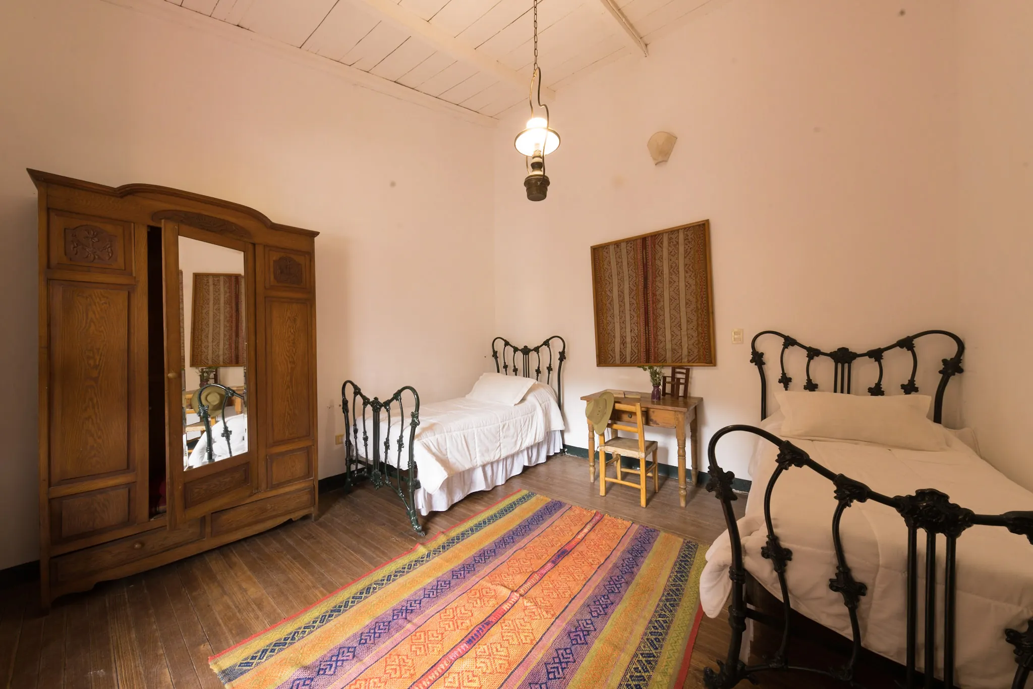 Cuarto con varias camas de hierro antiguas, ropero de madera y alfombra rayada de colores.