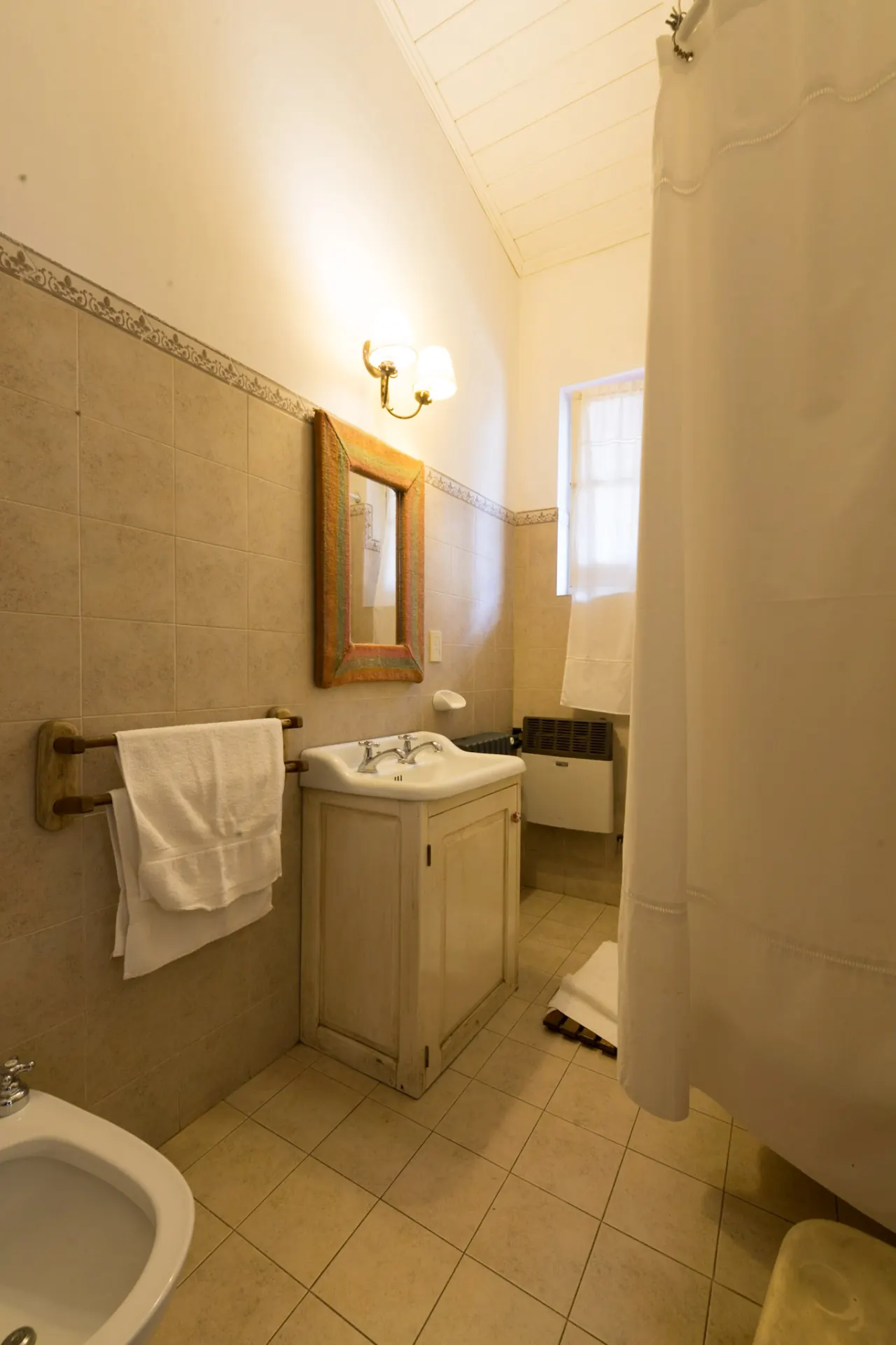 Baño revestido en cerámica clara, con lavatorio de madera y ventana alta.