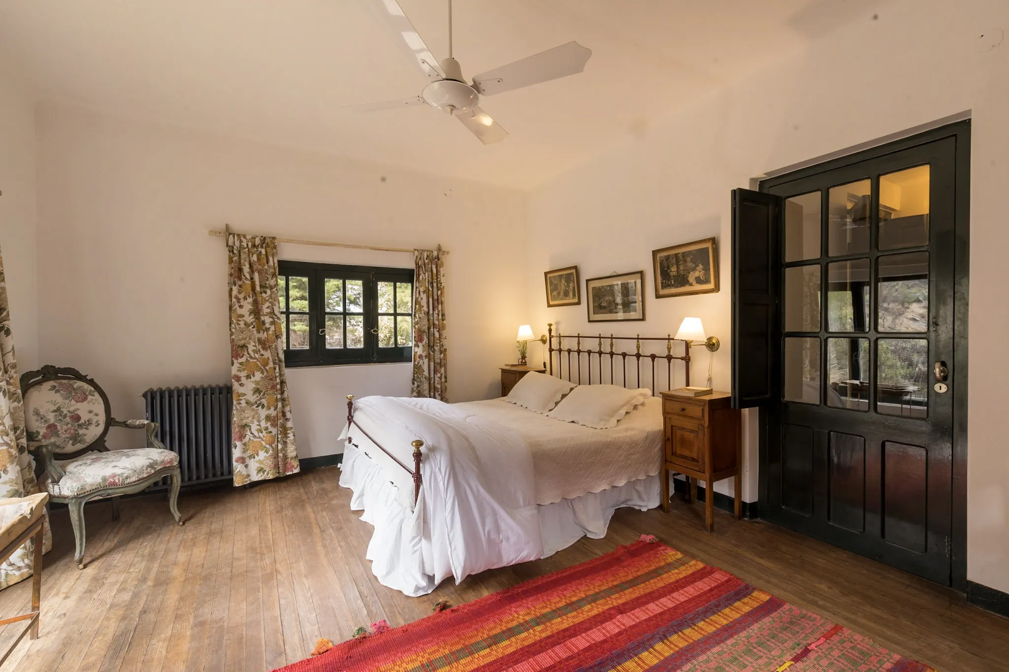 Habitación con cama de hierro, alfombra rayada de colores y puertas vidriadas verdes.