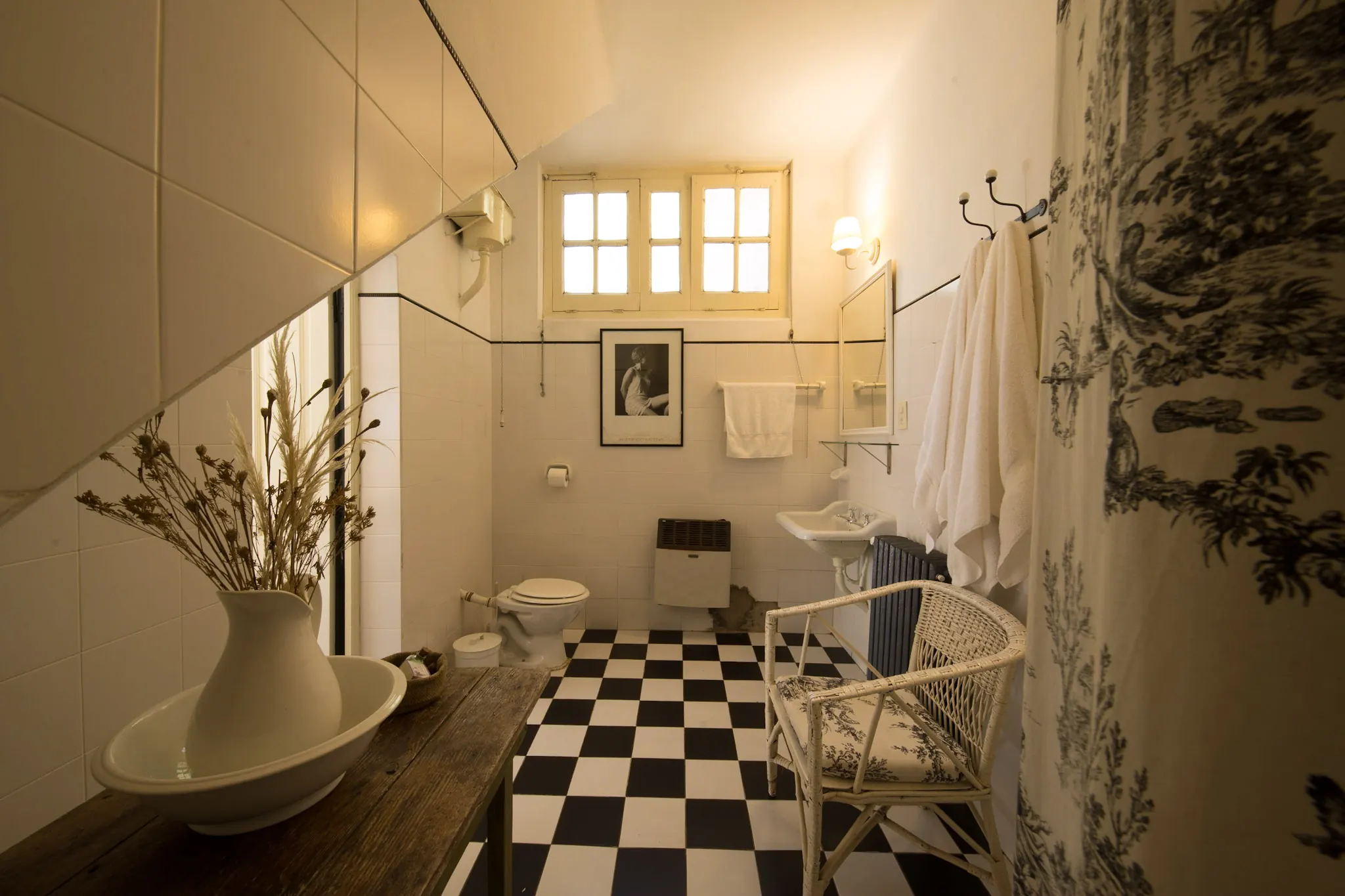 Baño amplio de piso ajedrezado blanco y negro, con ventana y una mesa de madera con jofaina.