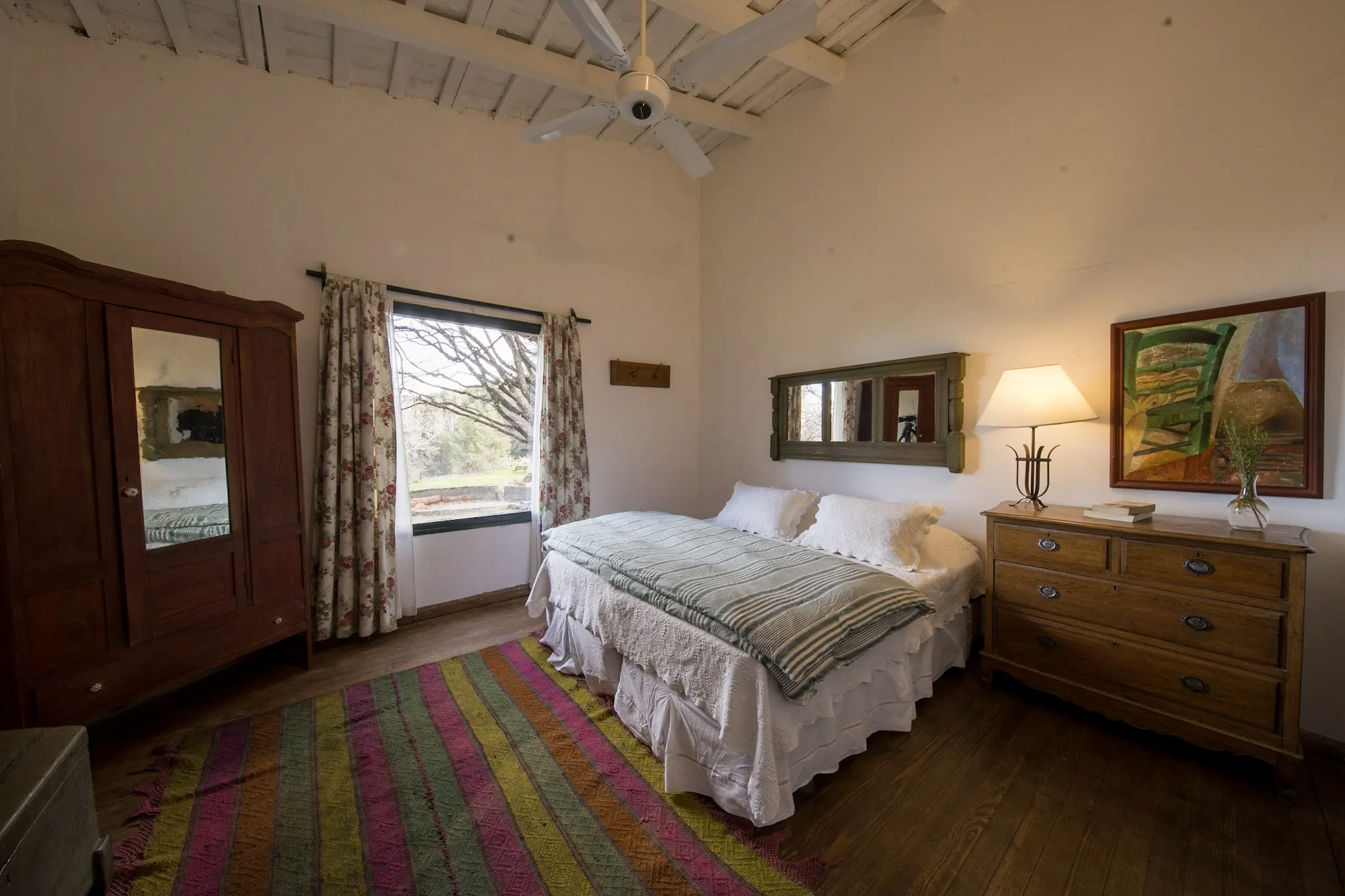 Habitación de techo alto blanqueado, con el ropero de madera, la ventana al patio y la cama con manta rayada.