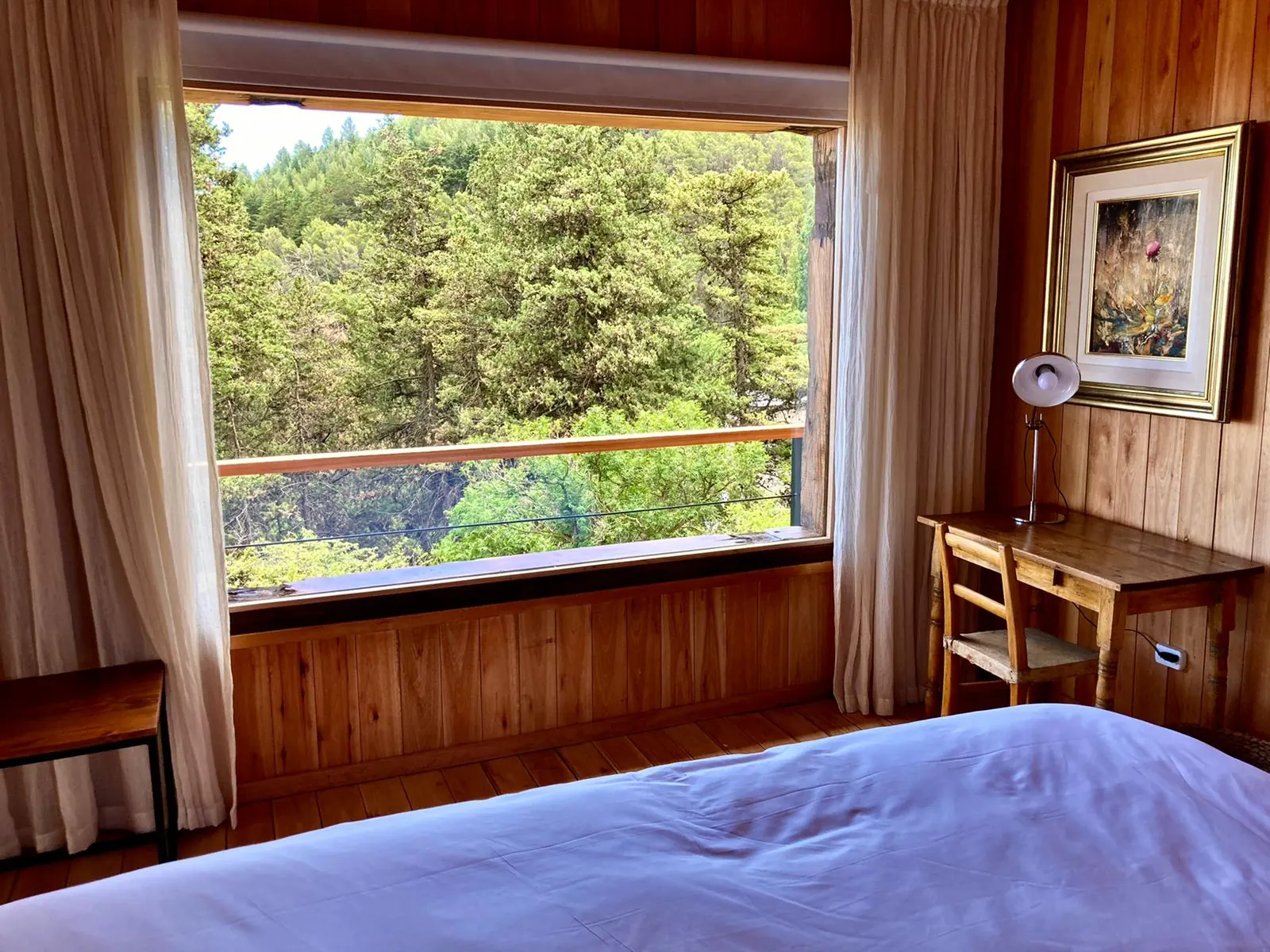 Interior de madera con la cama frente a un ventanal que da al bosque.