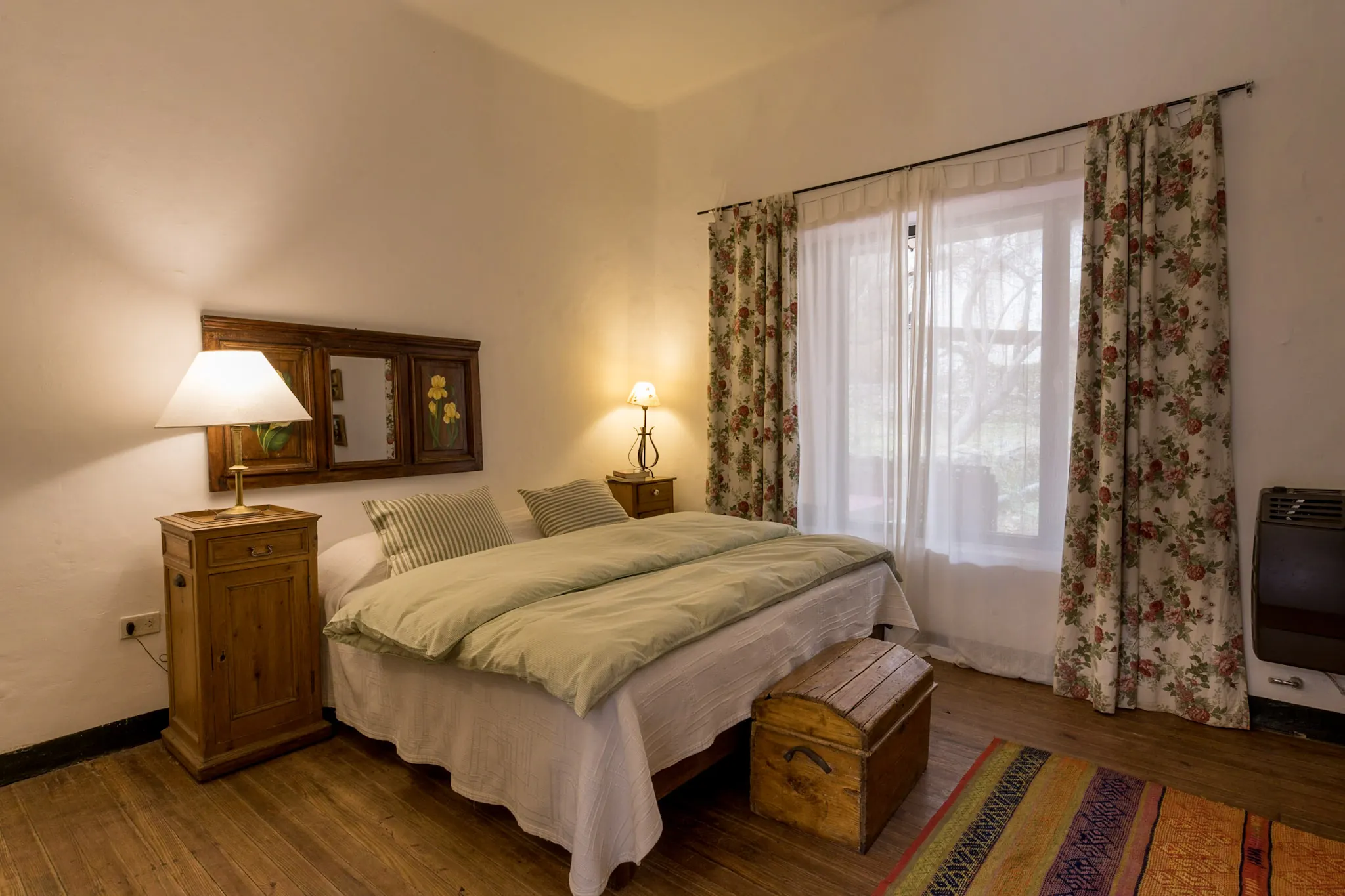 Habitación con cama matrimonial de acolchado verde, mesas de luz de madera y ventana con cortinas floreadas.