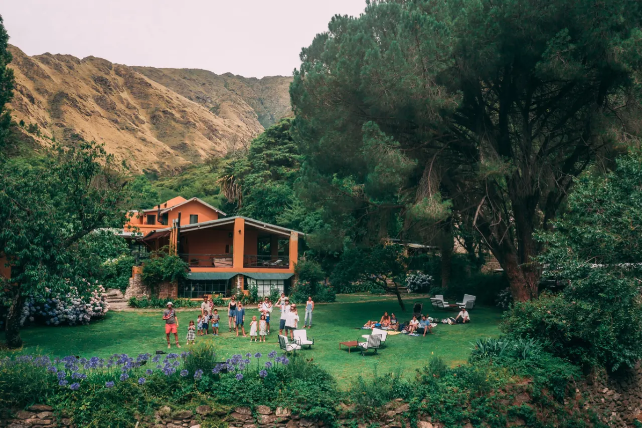 Familias con chicos repartidas por el jardín frente a la casa, unos parados sobre el césped y otros sentados en reposeras, con los cerros de la quebrada detrás.