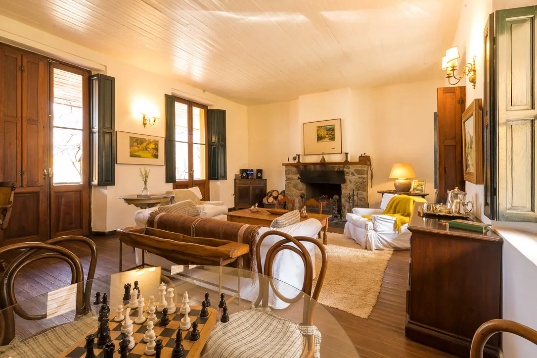 El living con el hogar a leña encendido al fondo, sillones con fundas blancas, un banco de madera y una mesa con un tablero de ajedrez.