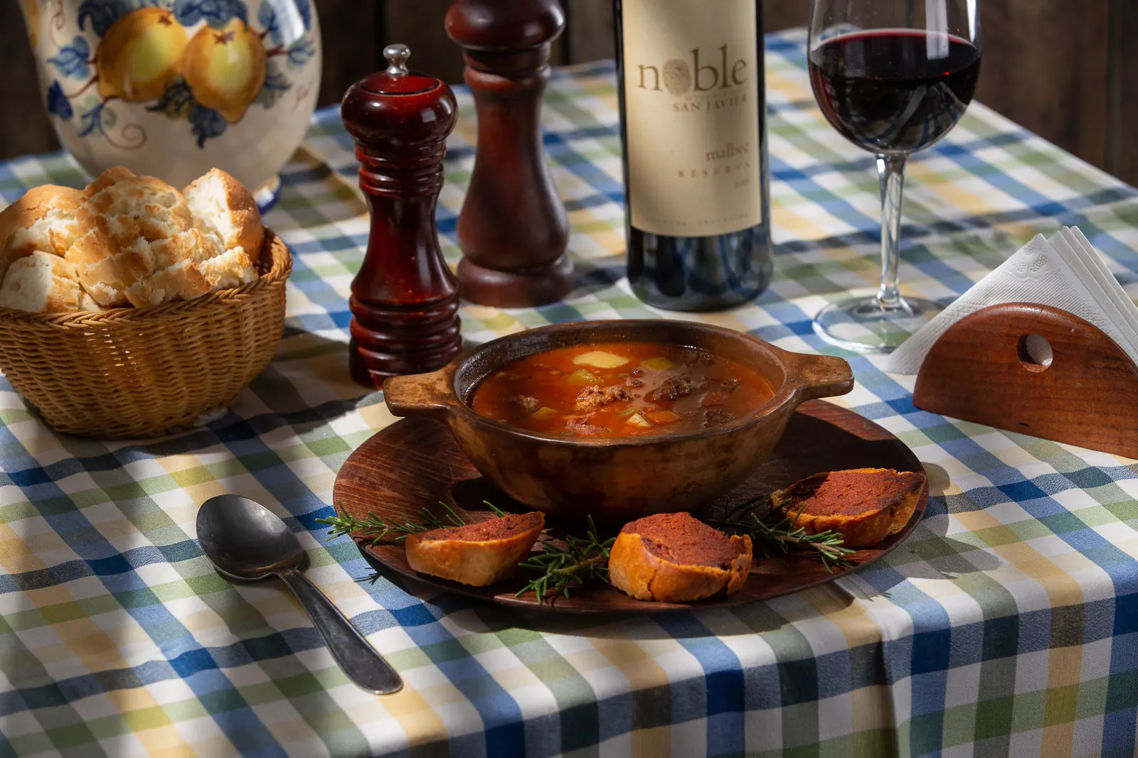 Un locro en cazuela de barro con tostadas de chorizo colorado, pan casero en canasto y un malbec de San Javier, sobre el mantel a cuadros.