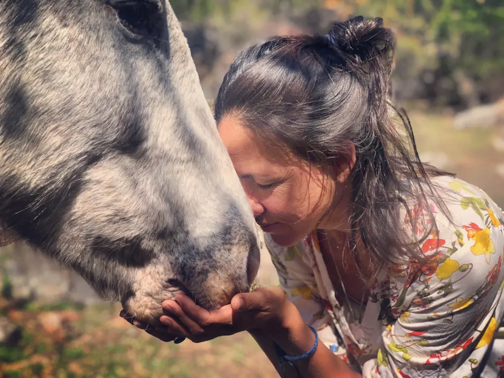 Una mujer con los ojos cerrados apoya la frente en el hocico de un caballo tordillo, sosteniéndolo con la mano.