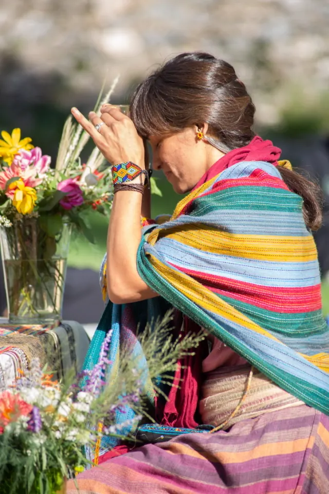 Una mujer envuelta en un rebozo de rayas de colores, con las manos juntas frente a la frente, al lado de un ramo de flores.