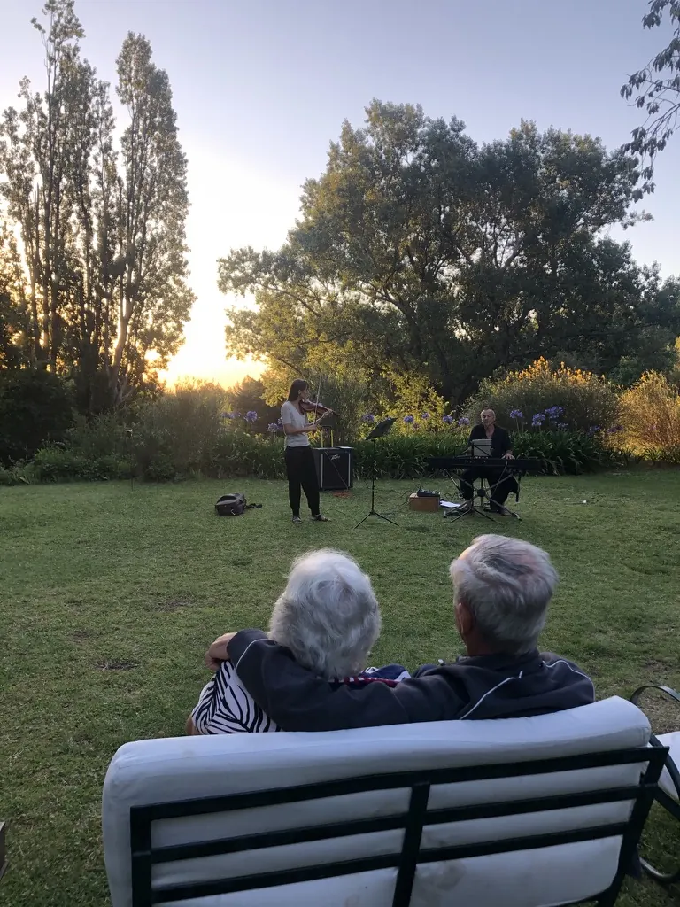 Una pareja mayor abrazada en un banco del jardín escucha a un violín y un teclado que tocan al atardecer, entre los agapantos.