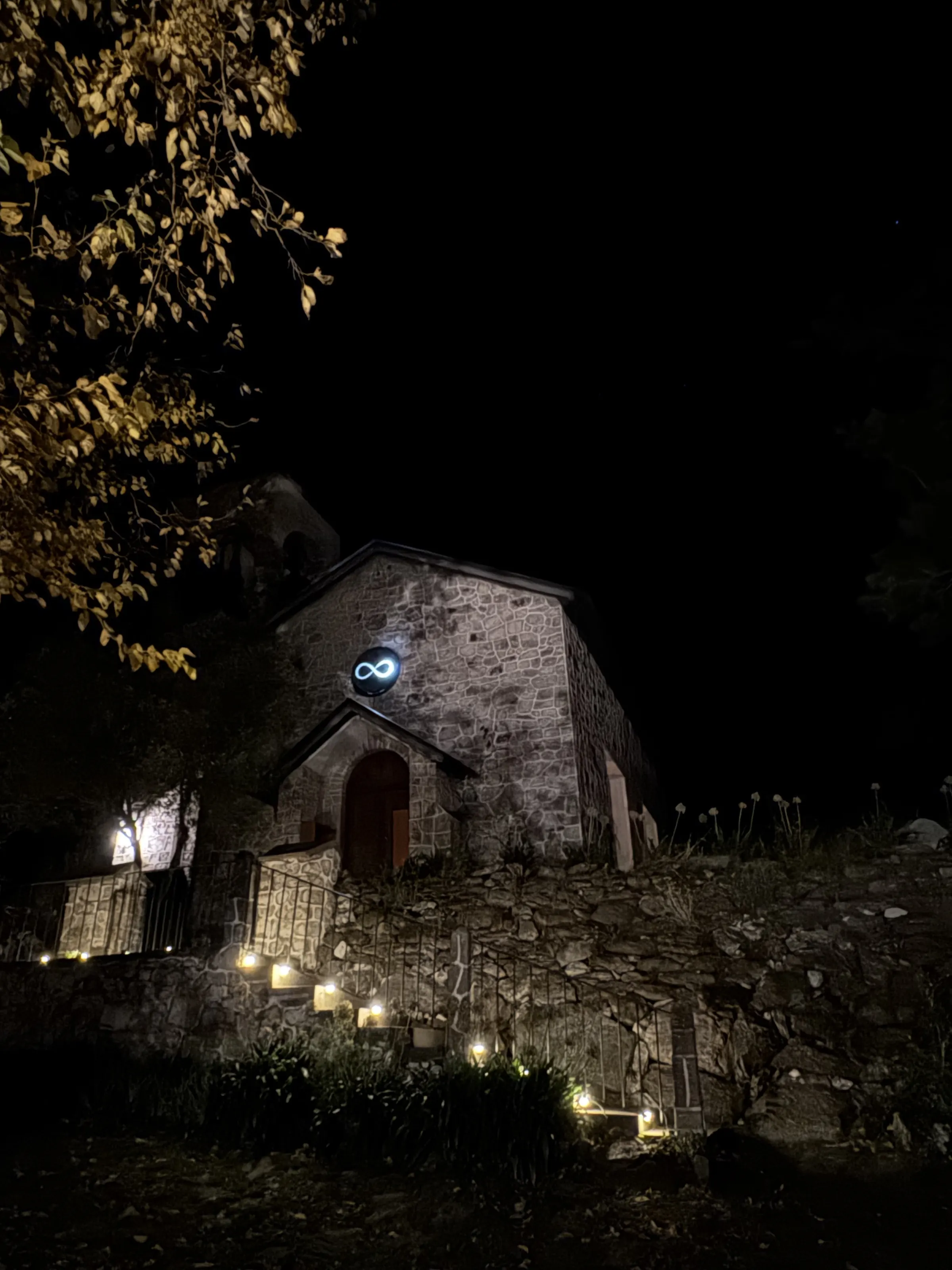 El oratorio de piedra de noche, con el símbolo del infinito iluminado sobre la puerta y velas encendidas en la escalera.
