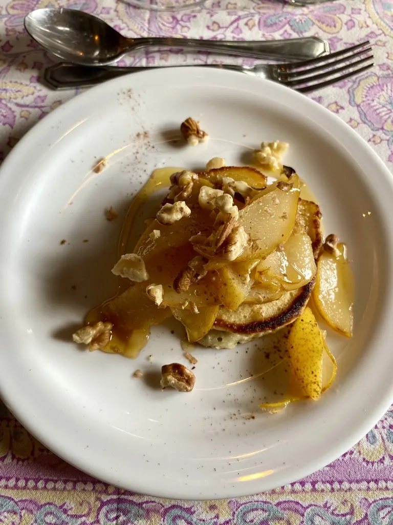 Un panqueque con peras caramelizadas, nueces y miel sobre un plato blanco, en un mantel estampado.
