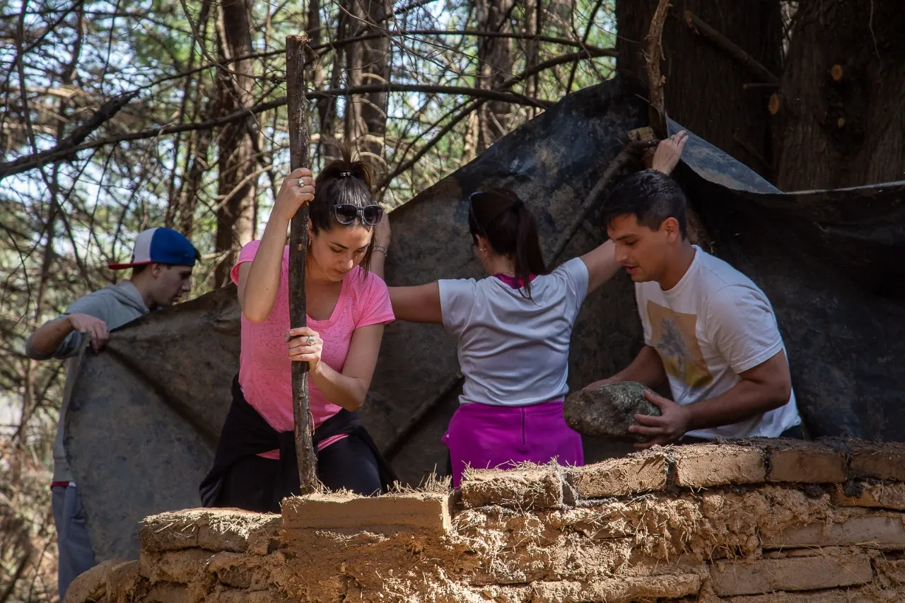 Cuatro personas levantando una pared de adobe entre los pinos: una apisona el barro con un palo y otra acerca una piedra.